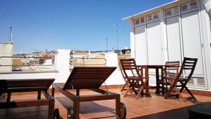 Eine Dachterrasse mit Blick auf die Stadt, mit Tisch und Stühlen, einem weißen Gebäude mit großen Fenstern und einem weißen Sonnenschirm.