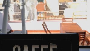 Blick durch ein Fenster, mit einem Schild, auf dem "CAFFÉ" steht, und einem Ausblick auf Sitzgelegenheiten und Tische im Freien.