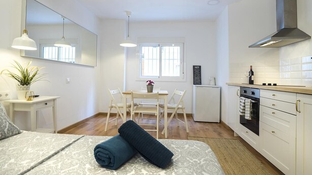 Une pièce compacte et bien éclairée, comprenant un lit, une table de chevet, un petit bureau, une table à manger avec des chaises, un coin cuisine et une grande fenêtre.