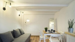 Ein modernes Wohnzimmer mit grauem Sofa, weißem Esstisch und Stühlen. Zwei Wandleuchten, ein kleiner Topfpflanze und ein Spiegel sind an der Wand angebracht.