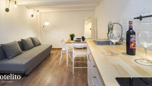 Ein kompakter Wohnbereich mit Sofa, Esstisch, Stühlen, einer kleinen Küchenzeile mit Spüle sowie Weingläsern und einer Weinflasche.