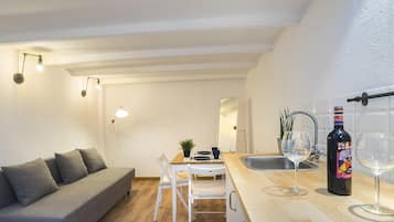 A compact living space with a sofa, dining table, chairs, a small kitchen area with a sink and wine glasses, and a bottle of wine.