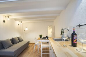 A compact living space with a sofa, dining table, chairs, a small kitchen area with a sink and wine glasses, and a bottle of wine.