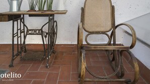 Una silla rústica con respaldo tejido y una mesa de máquina de coser de metal con una jarra blanca y plantas verdes encima.
