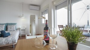 Una sala de estar moderna con sofá, una mesa de comedor con copas de vino y una botella, una planta en maceta y un balcón con vistas.