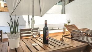 Una mesa de madera con una copa de vino y una botella, una planta en maceta y un paraguas blanco en un balcón.