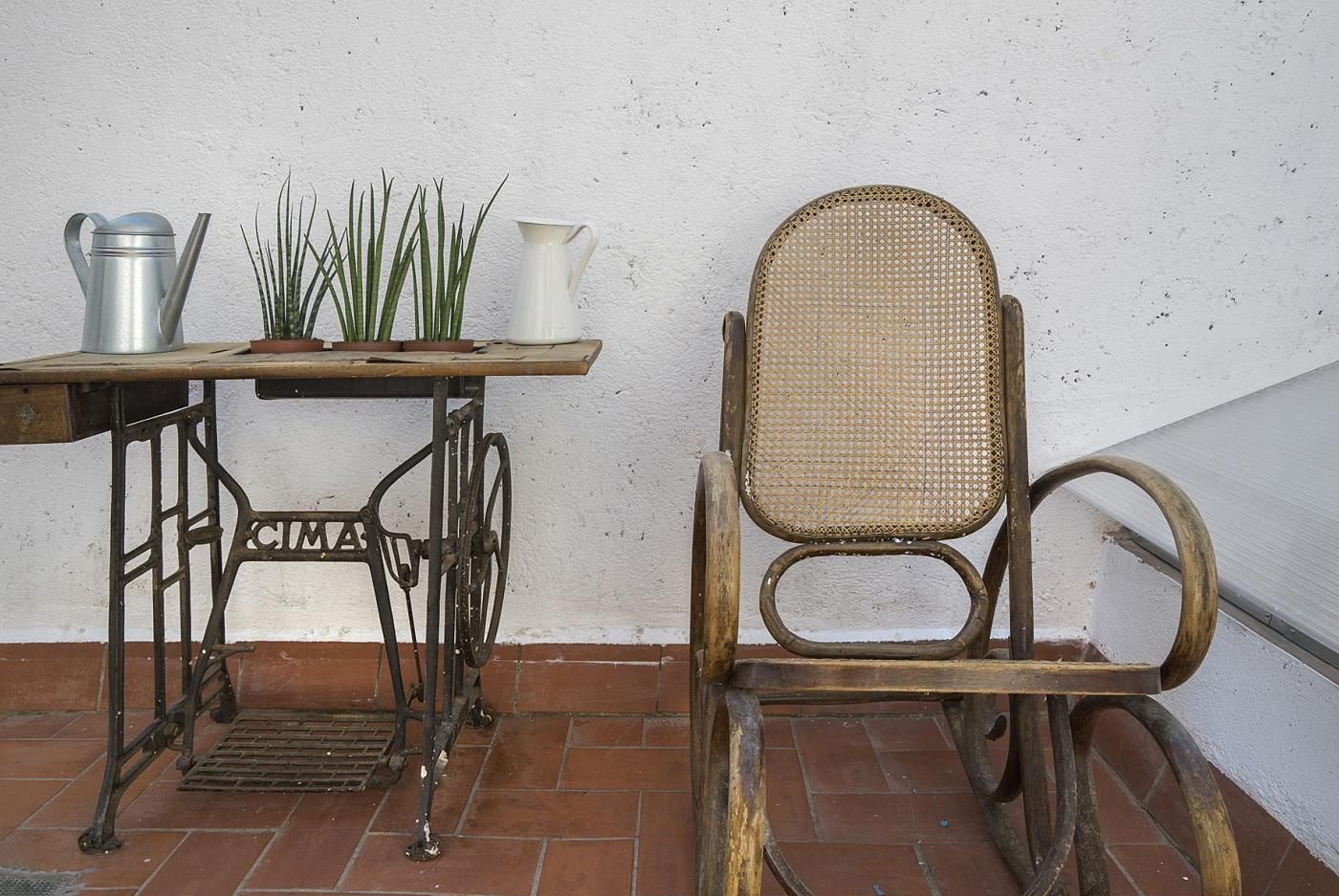 Una silla rústica con respaldo tejido y una mesa de máquina de coser de metal con una jarra blanca y plantas verdes encima.