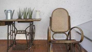 Una silla rústica con respaldo tejido y una mesa de máquina de coser de metal con una jarra blanca y plantas verdes encima.