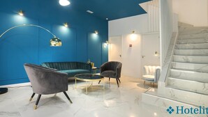 Um saguão moderno de hotel com uma parede azul de destaque, uma mesa de centro redonda, duas poltronas e uma escadaria de mármore.