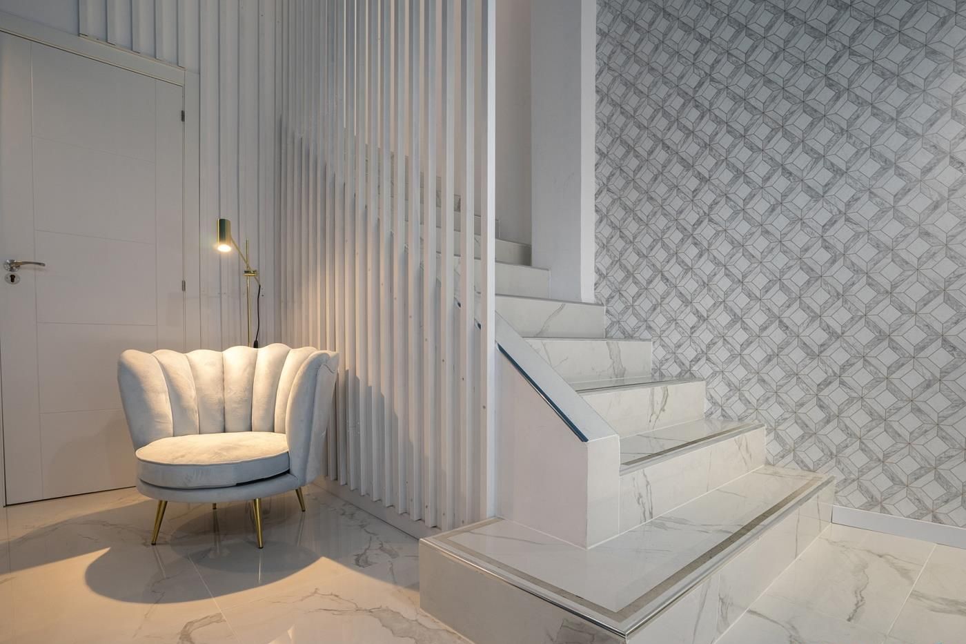 Ein modernes Interieur mit einem weißen, kuscheligen Sessel, einer Treppe mit geometrisch gemusterten Wänden und einem Marmorboden.