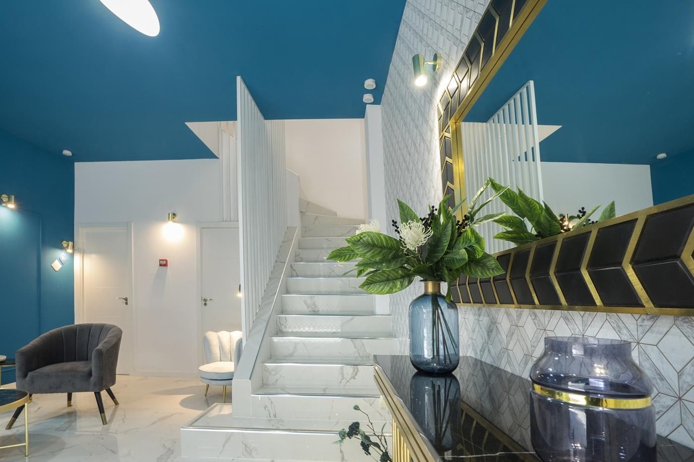 Un lobby moderno de hotel con una escalera de mármol, una zona de estar y un espejo grande con detalles decorativos.