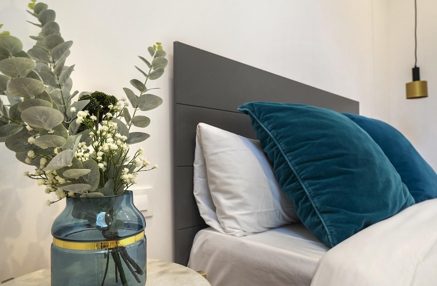 A bedroom with a bed, a nightstand, a vase with flowers, and a wall-mounted lamp.