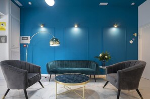 Ein Zimmer mit blauer Akzentwand, einem dunkelgrünen Sofa, zwei grauen Sesseln, einem runden Marmor-Couchtisch und einer Stehlampe.