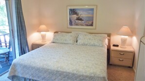 A neatly made bed with a quilted cover, two bedside tables with lamps, a framed picture on the wall, and a view of the outdoors through a window. - Ariel East Duplex 3 bedrooms, 2.5 bathrooms (Jekyll Island)