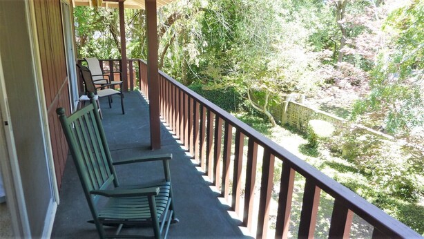 A covered balcony with a wooden railing, two green chairs, and a view of a garden with trees and a fence. - Ariel East Duplex 3 bedrooms, 2.5 bathrooms (Jekyll Island)