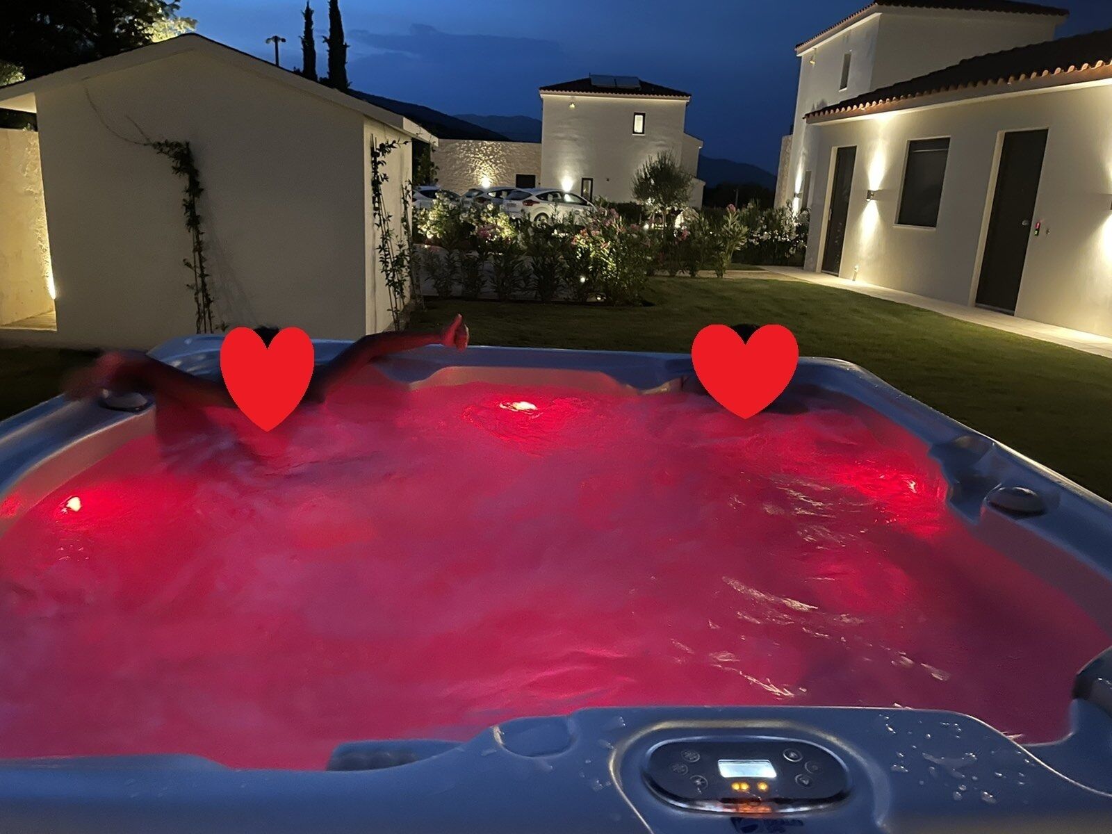 A hot tub with red lighting, surrounded by a well-maintained garden and a large house in the background.