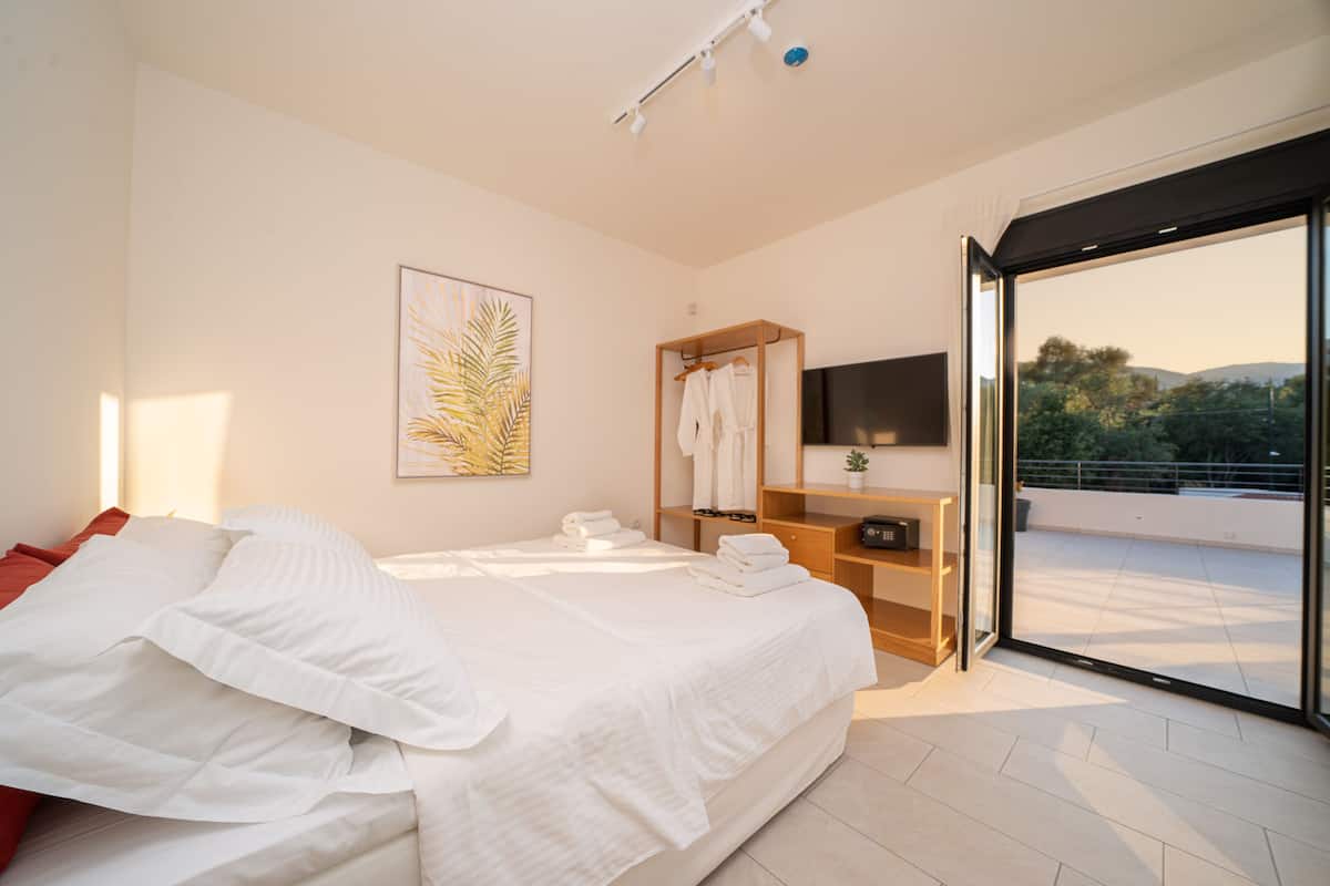 Une chambre moderne avec un grand lit, une commode en bois, un téléviseur à écran plat et une porte-fenêtre coulissante donnant sur un balcon avec vue sur la ville.
