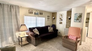 Ein Wohnzimmer mit einem dunkelbraunen Sofa, einem hellbraunen Sessel und einem kleinen Tisch mit einer Lampe. An den Wänden hängen gerahmte Bilder und ein Fenster mit Jalousien.