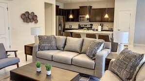 Un salon moderne avec un canapé beige, deux tables d’appoint, une table basse agrémentée de plantes, et une cuisine en arrière-plan.