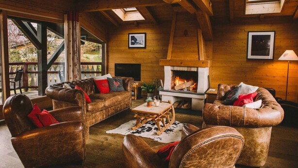 A cozy log cabin interior with leather sofas, a fireplace, and large windows overlooking a garden.