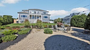 Een tweelaags huis met een overdekte veranda, een omheinde tuin en een zandgedeelte met tuinmeubilair.