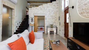 Ein modernes Wohnzimmer mit Steinwand, einer Treppe, einer Couch mit orangefarbenen Kissen, einem Fernseher und einem kleinen Tisch mit einem Weinglas.