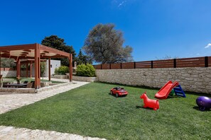 A patio with a pergola, a stone wall, a grassy lawn, a playground with a slide, and a tree.