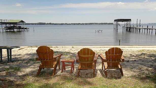 Uitzicht op een meer met drie houten stoelen en een kleine tafel in het zand, met een kalm wateroppervlak en land in de verte.