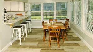 Een eetgedeelte met een houten tafel en stoelen, een keukenbar met krukken, en grote ramen met zonwering.