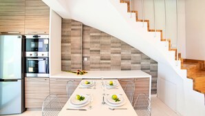 A modern kitchen with a built-in oven and refrigerator, a dining area with a white table set for four, and a wooden staircase with a unique design.