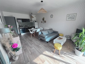 Un salon moderne avec un canapé gris, une table à manger avec des chaises, une cuisine en arrière-plan et une plante en pot.