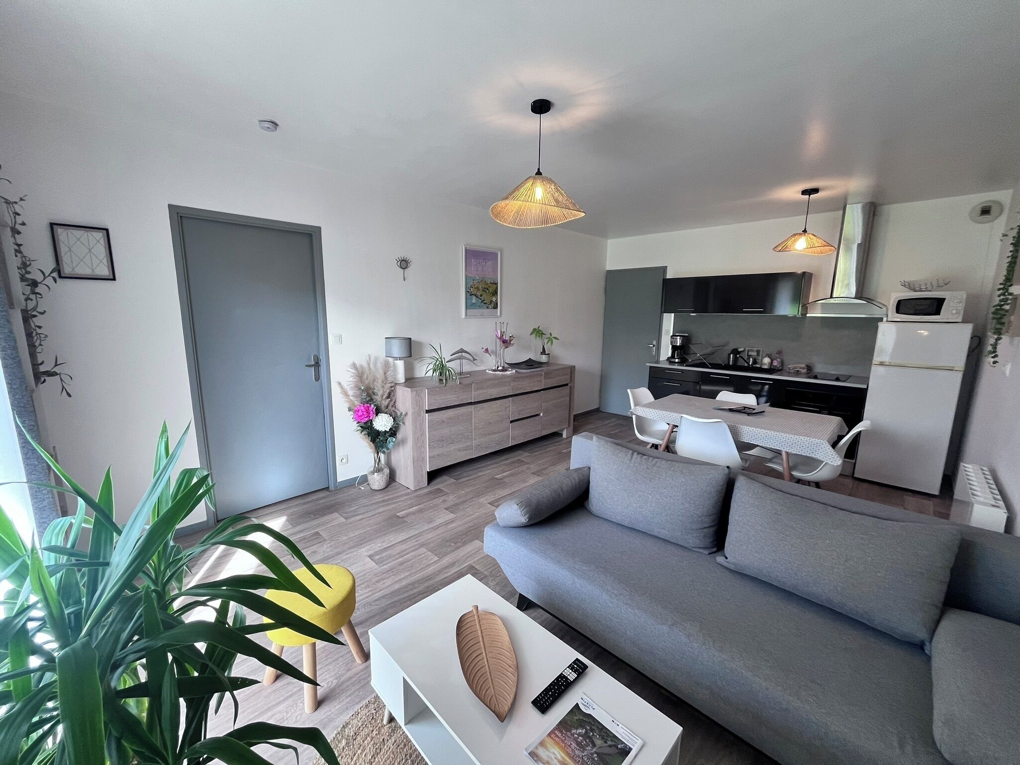 Una sala moderna con un sofá gris, una mesa de centro y un comedor con una mesa pequeña y sillas. Hay una zona de cocina con microondas y un refrigerador.