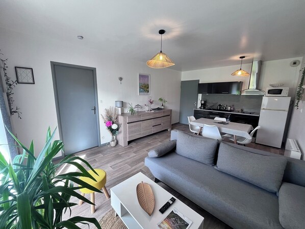 A modern living room with a grey sofa, a coffee table, and a dining area with a small table and chairs. There is a kitchen area with a microwave and a refrigerator.