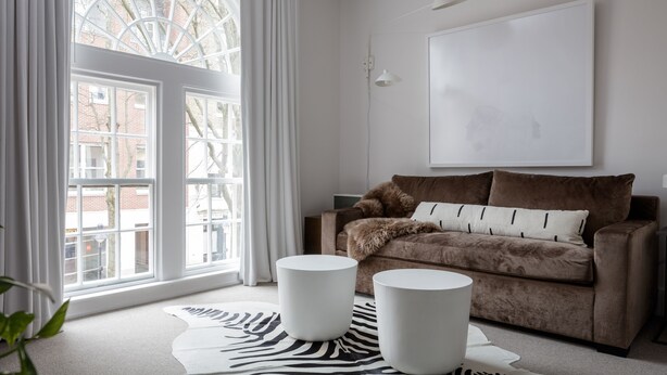 Un salon moderne avec une grande fenêtre, un canapé marron, deux petites tables d’appoint blanches et un tapis à motifs zébrés.