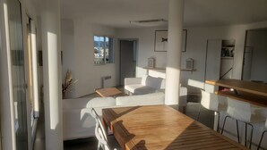 Ett modernt vardagsrum med ett matbord i trä, vita soffor och utsikt över staden genom stora fönster.