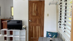 A compact living space with a wooden door, a TV on a white shelf, a small table with a remote, and a patterned rug. - Bak A  Yaad Cottage Lupine Cottage (Negril)