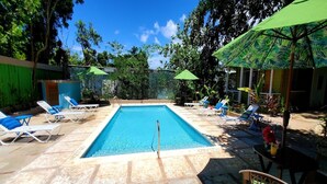 A rectangular pool with a ladder, surrounded by lounge chairs and umbrellas, set in a tiled area with trees in the background. - Bak A  Yaad Cottage Lupine Cottage (Negril)