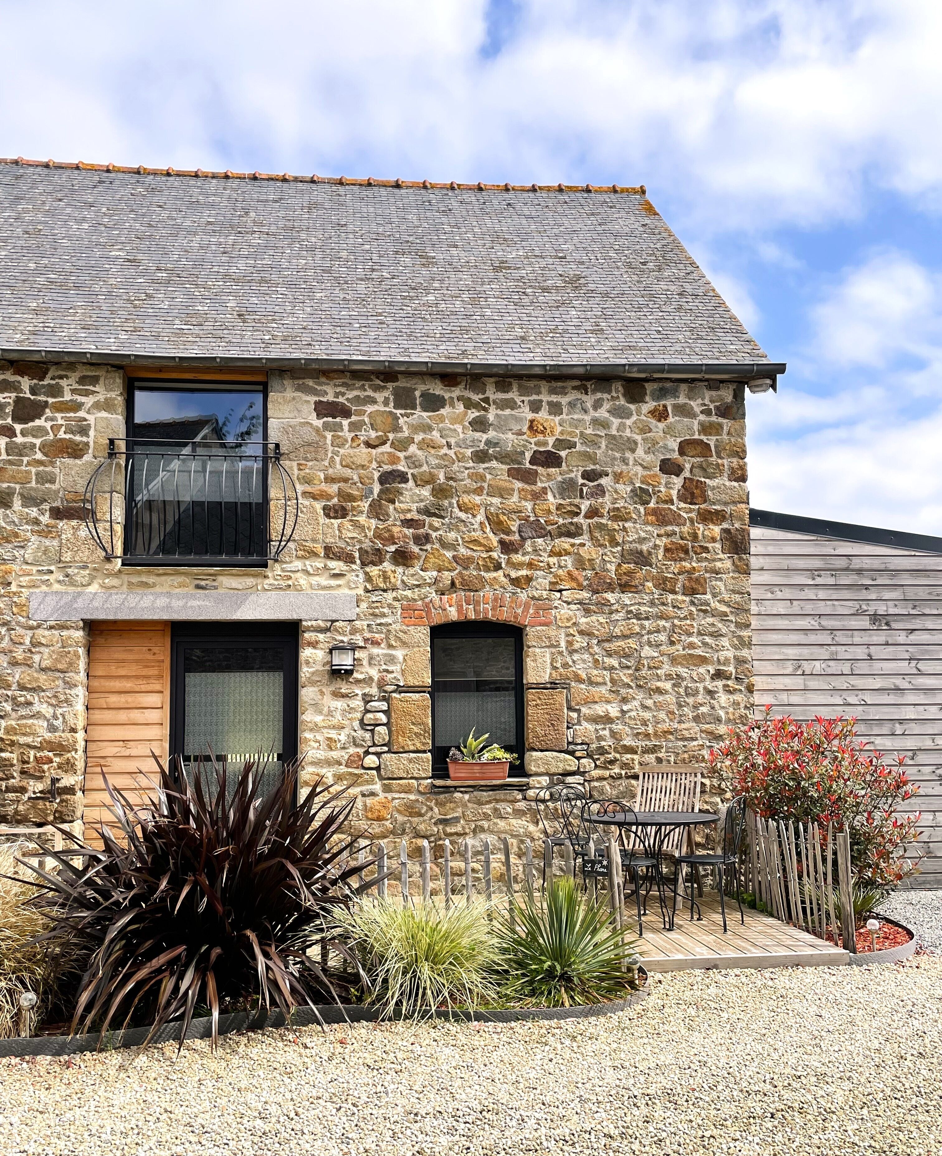 A stone building with a wooden door, a window with a metal railing, and a small garden area with plants and a wooden fence.