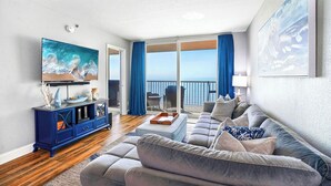 A living room with a large flat-screen TV, a blue entertainment unit, a grey sofa, and a balcony with a view of the sea. - Beachfront  Paradise (Indian Shores)
