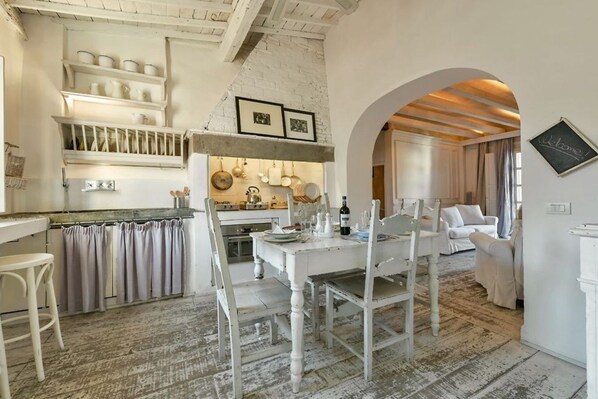 A rustic kitchen with white cabinetry, a dining table set with chairs, and a view into a living area with a white sofa.