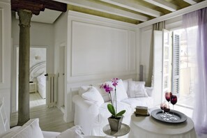 A white room with a bed, a white sofa, a round table with a tray and glasses, and a window with shutters.