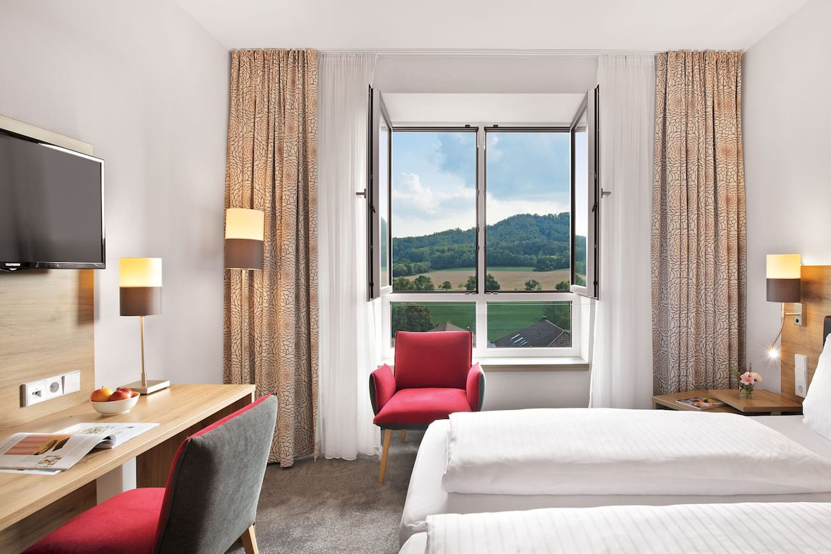 A hotel room with a bed, a desk, a chair, a TV, and a window with a view of a green landscape.