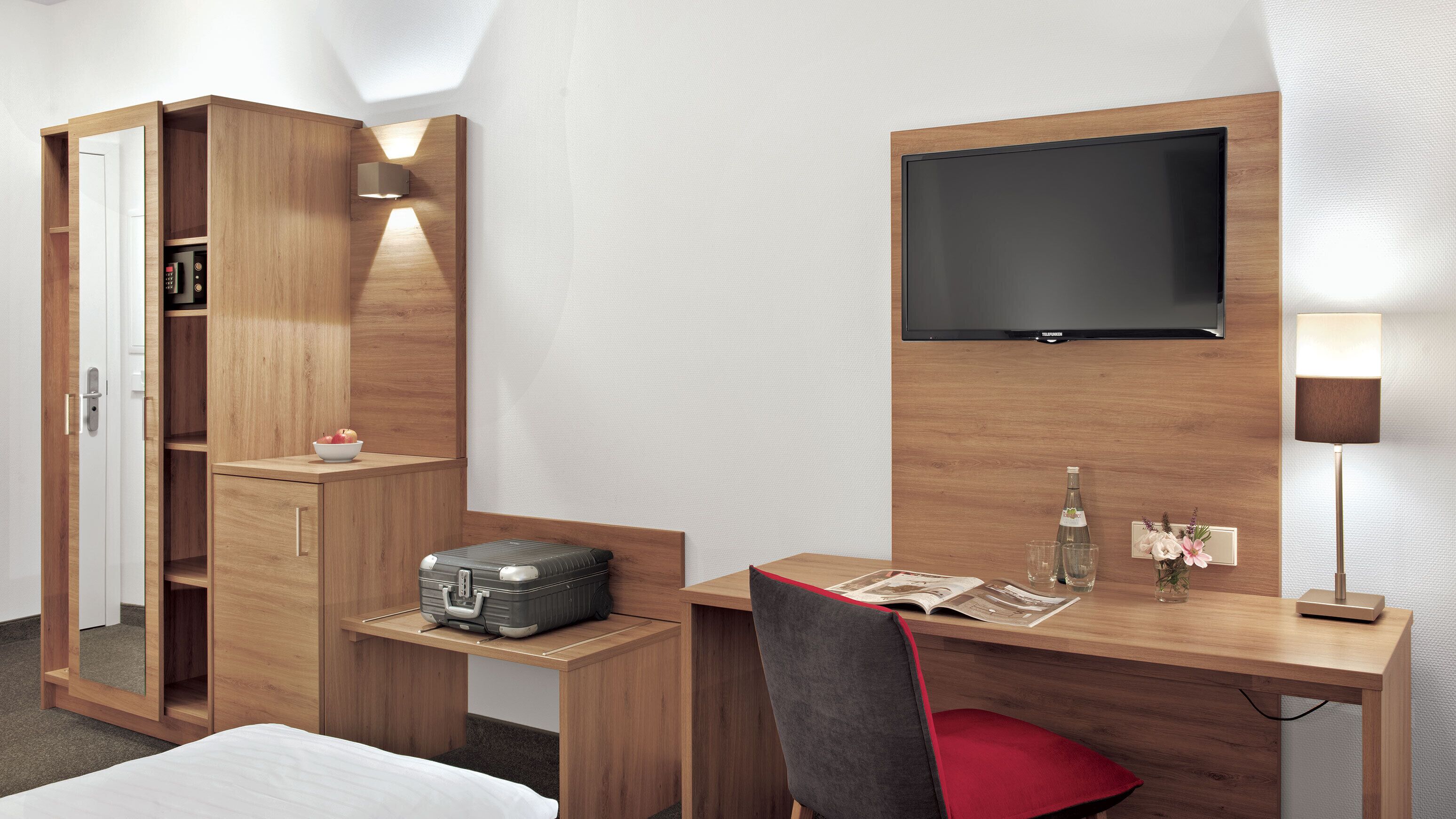A hotel room with a wooden desk, a flat-screen TV, a bedside table with a lamp, a wardrobe, and a bed.