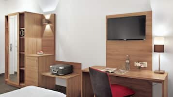 A hotel room with a wooden desk, a flat-screen TV, a bedside table with a lamp, a wardrobe, and a bed.