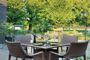 Un conjunto de mesa y sillas de mimbre con un bol de fruta, copas de vino y cubiertos en un balcón con vistas a exuberante vegetación.