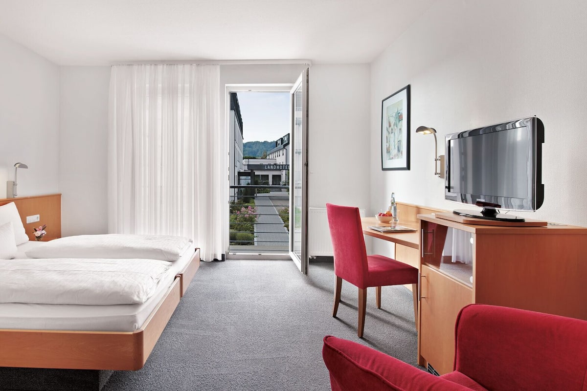 A hotel room with a bed, a desk with a TV, a chair, and a view of a building outside.