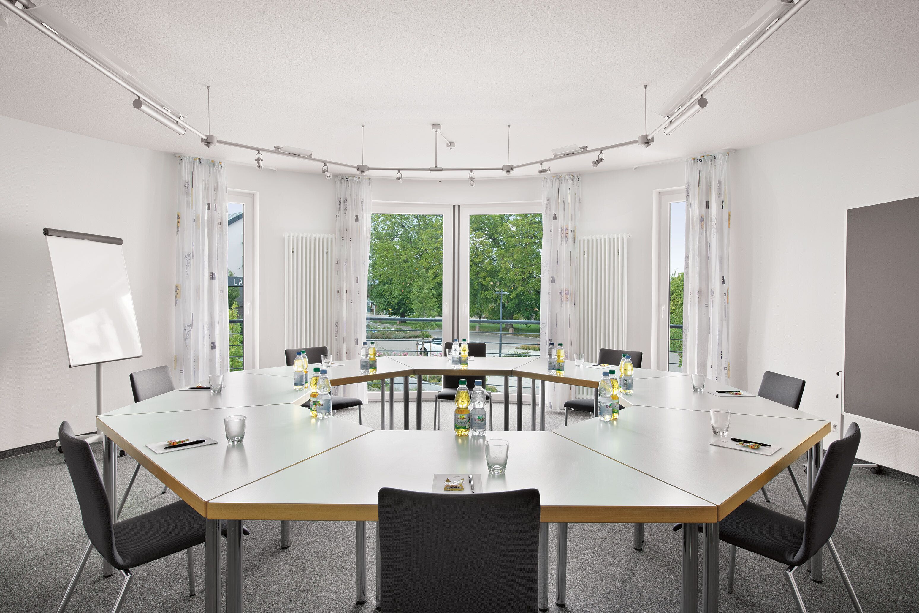 A conference room with a large oval table, black chairs, a whiteboard, and large windows with curtains.