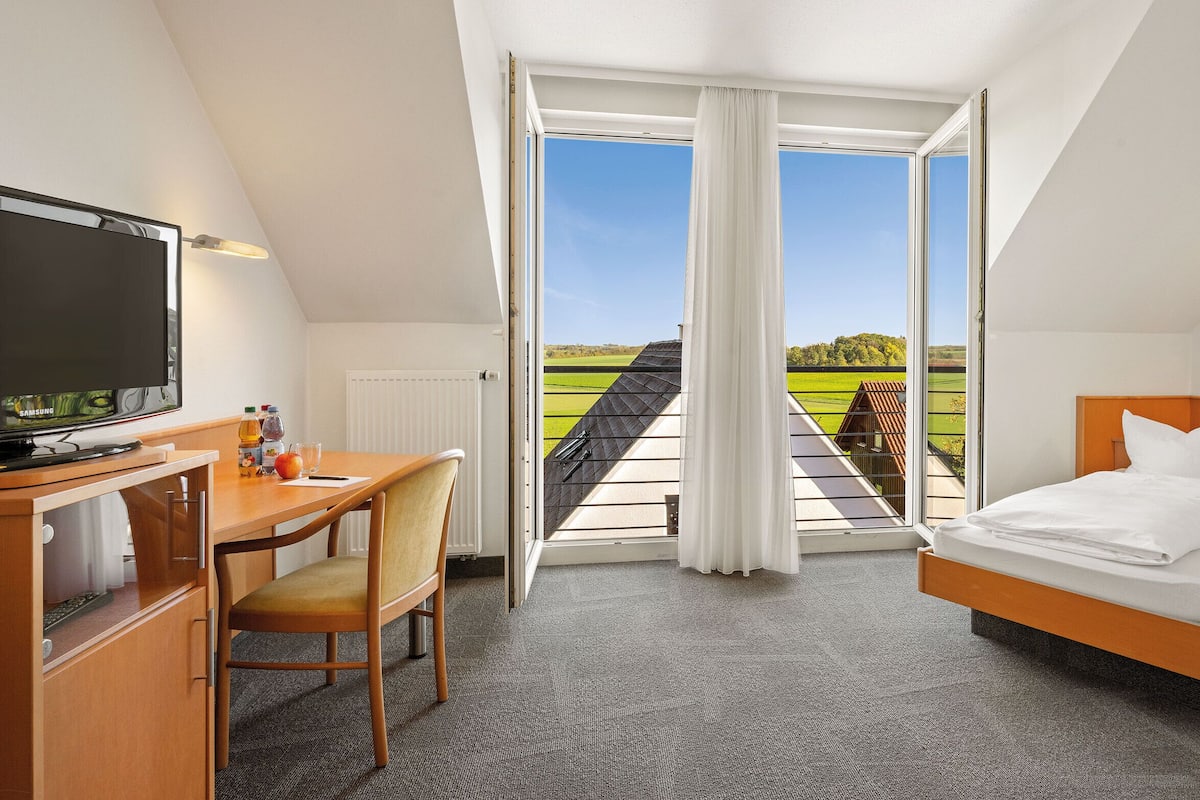 A hotel room with a bed, desk, chair, television, and a view of a green landscape through a large window.
