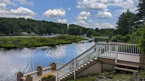 Une terrasse en bois, dotée d’une rambarde, mène à un quai surplombant une rivière paisible, le tout étant entouré de verdure et d’arbres.