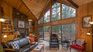 A spacious living area with a leather sofa, a red chair, and a wooden coffee table. There is a stone wall with framed pictures and a fireplace. Large windows offer a view of the surrounding forest. - Mink #8 Close to SHARC and 8 SHARC Passes! Close to Village! (Sunriver)